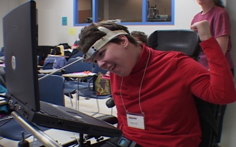 車いすの若い女性がヘッドスティックを使ってコンピュータを使っている。